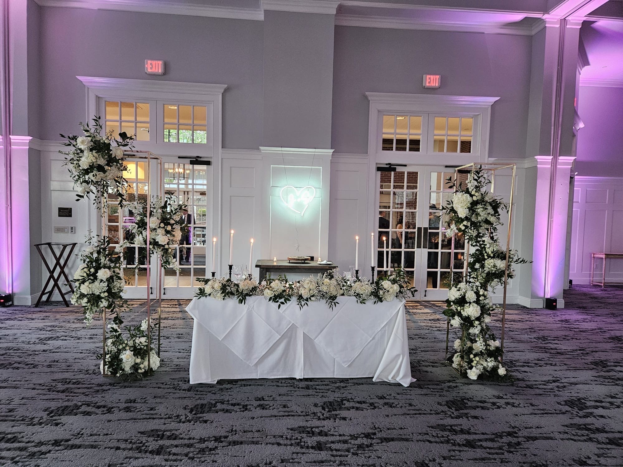 Sweetheart table framed by two gold arbors of white roses, hydrangea and greenery under soft violet lighting