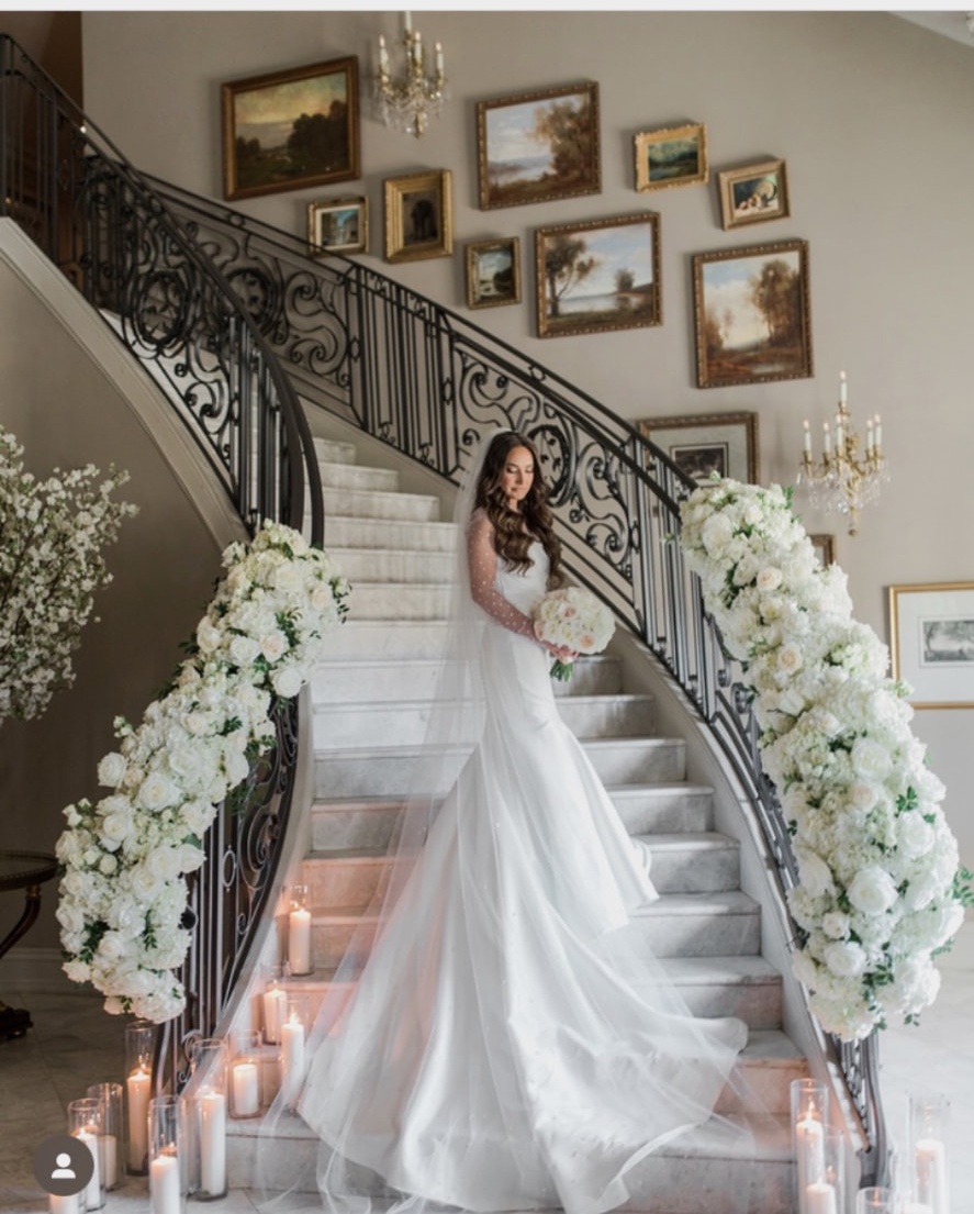 Bride on a marble staircase framed by cascading white rose and hydrangea installations and pillar candles