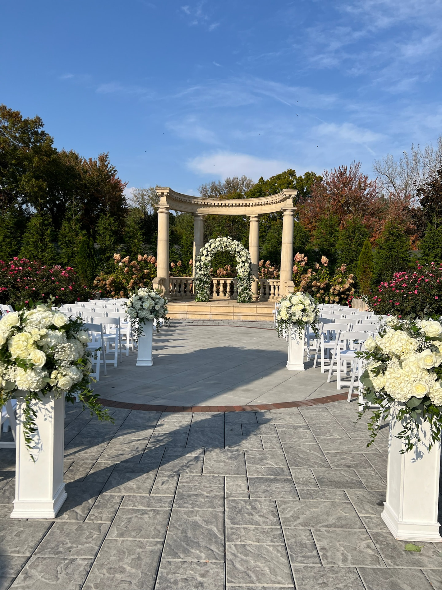 Outdoor garden ceremony with a white floral arch beneath a stone pergola and aisle pedestal arrangements