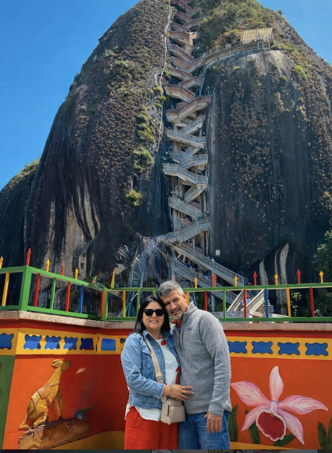 Yumila with her husband while travelling, standing in front of a brightly painted railing and a towering rock formation