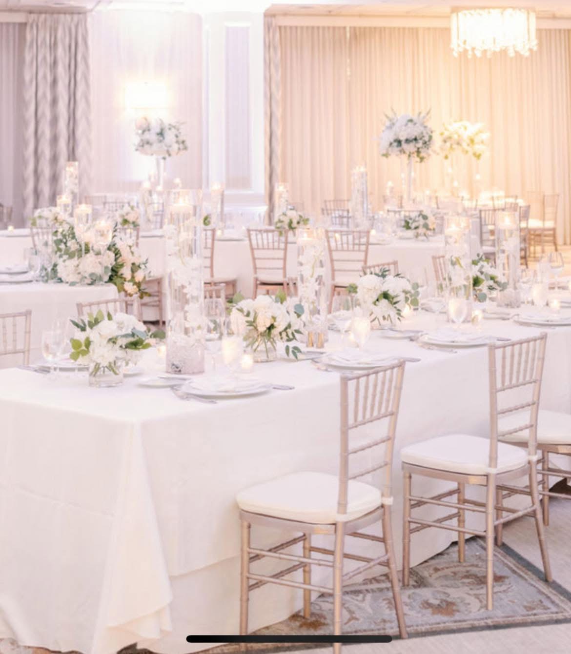 White ballroom reception with blush chiavari chairs, white floral clusters, and orchid-filled candle cylinders
