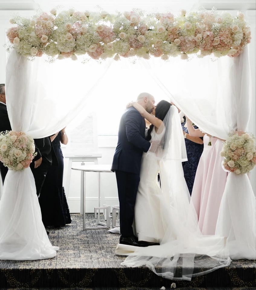 Couple kissing beneath a blush and ivory chuppah draped in sheer white fabric