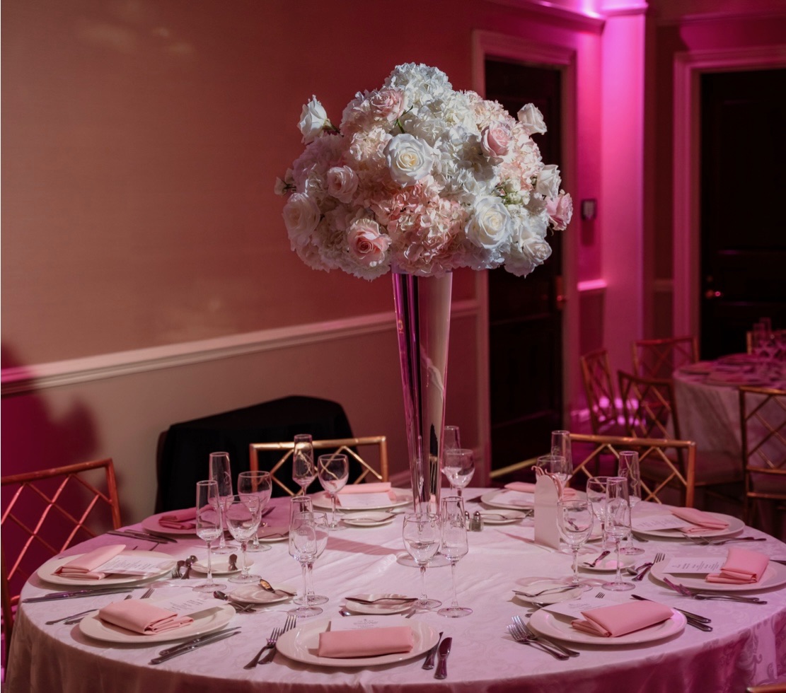 Tall trumpet vase centerpiece of white and blush roses and hydrangea in a pink-lit ballroom