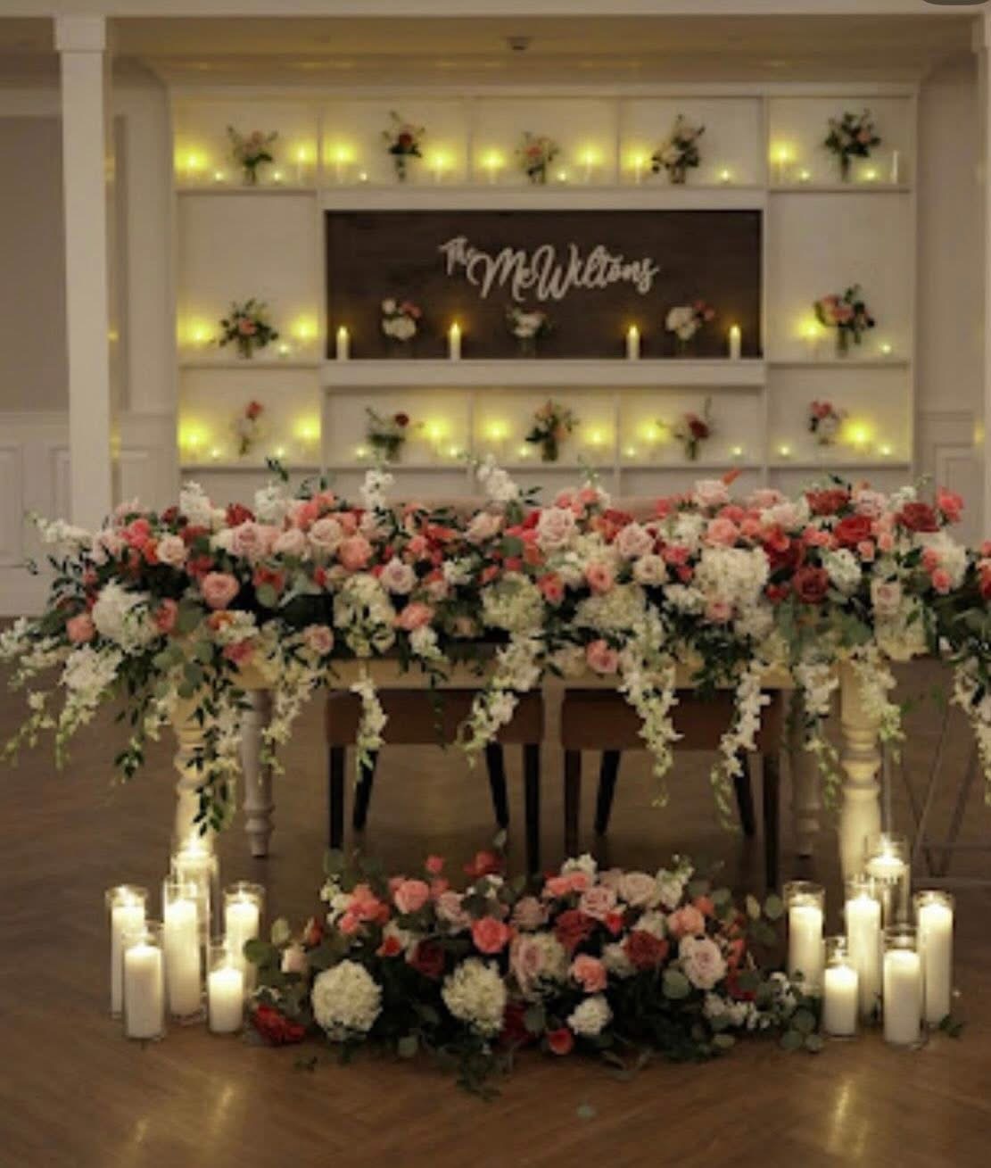 Sweetheart table draped in blush, coral, and burgundy blooms beneath a lit display wall
