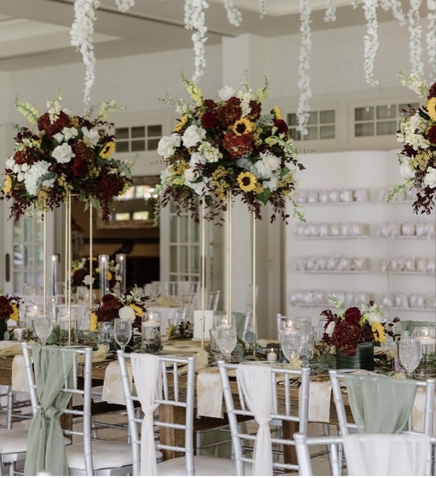 Long harvest table with tall burgundy, sunflower and cream centerpieces on gold stands beneath hanging white florals