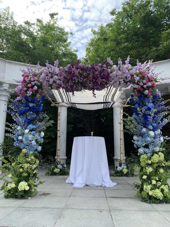 Garden chuppah with birch poles draped in purple, lavender and blue florals over green hydrangea