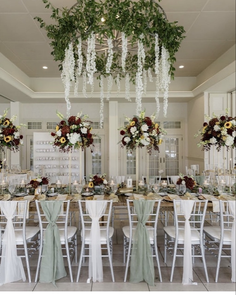 Ballroom with a greenery chandelier of white wisteria over tall burgundy, sunflower and white centerpieces