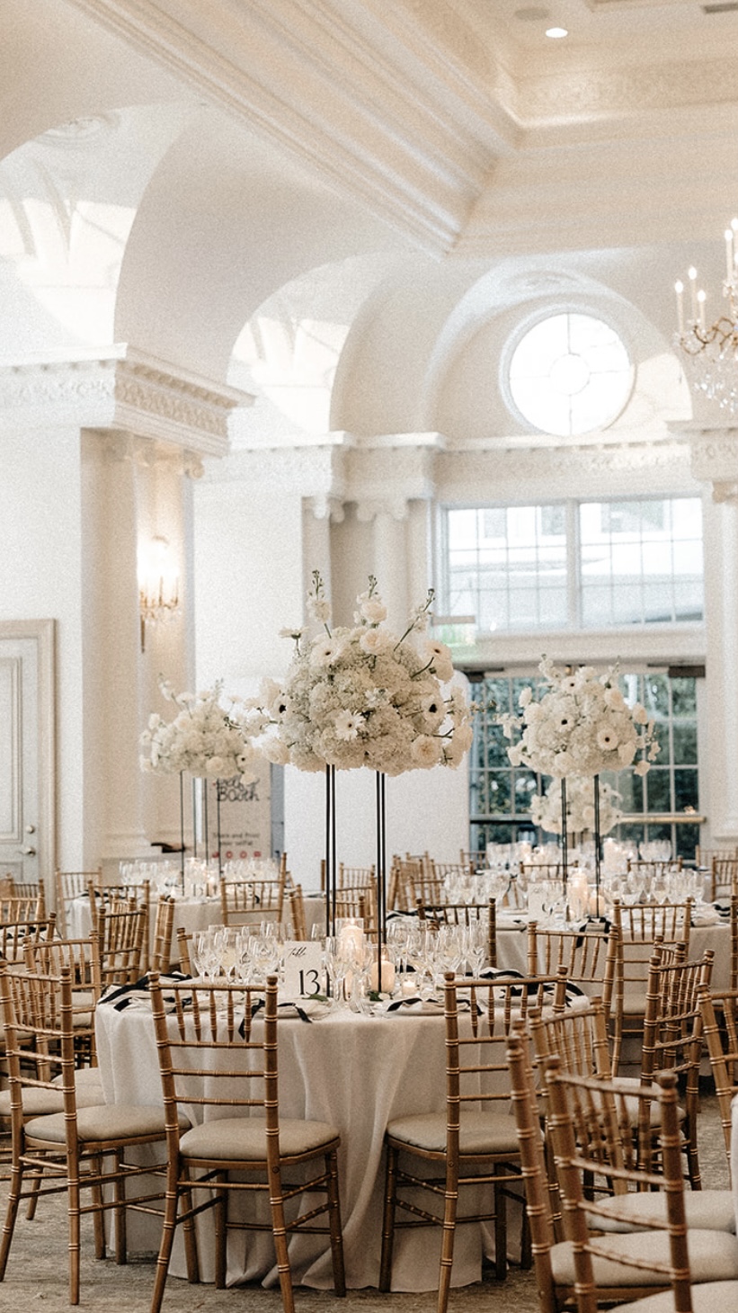 Bright ballroom of round tables with tall all-white anemone and ranunculus centerpieces on black stands