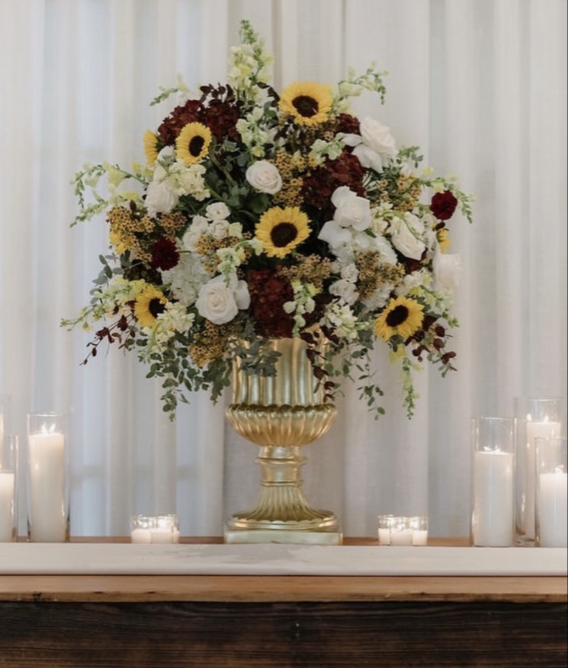 Gold urn overflowing with sunflowers, burgundy hydrangea and white roses above pillar candles
