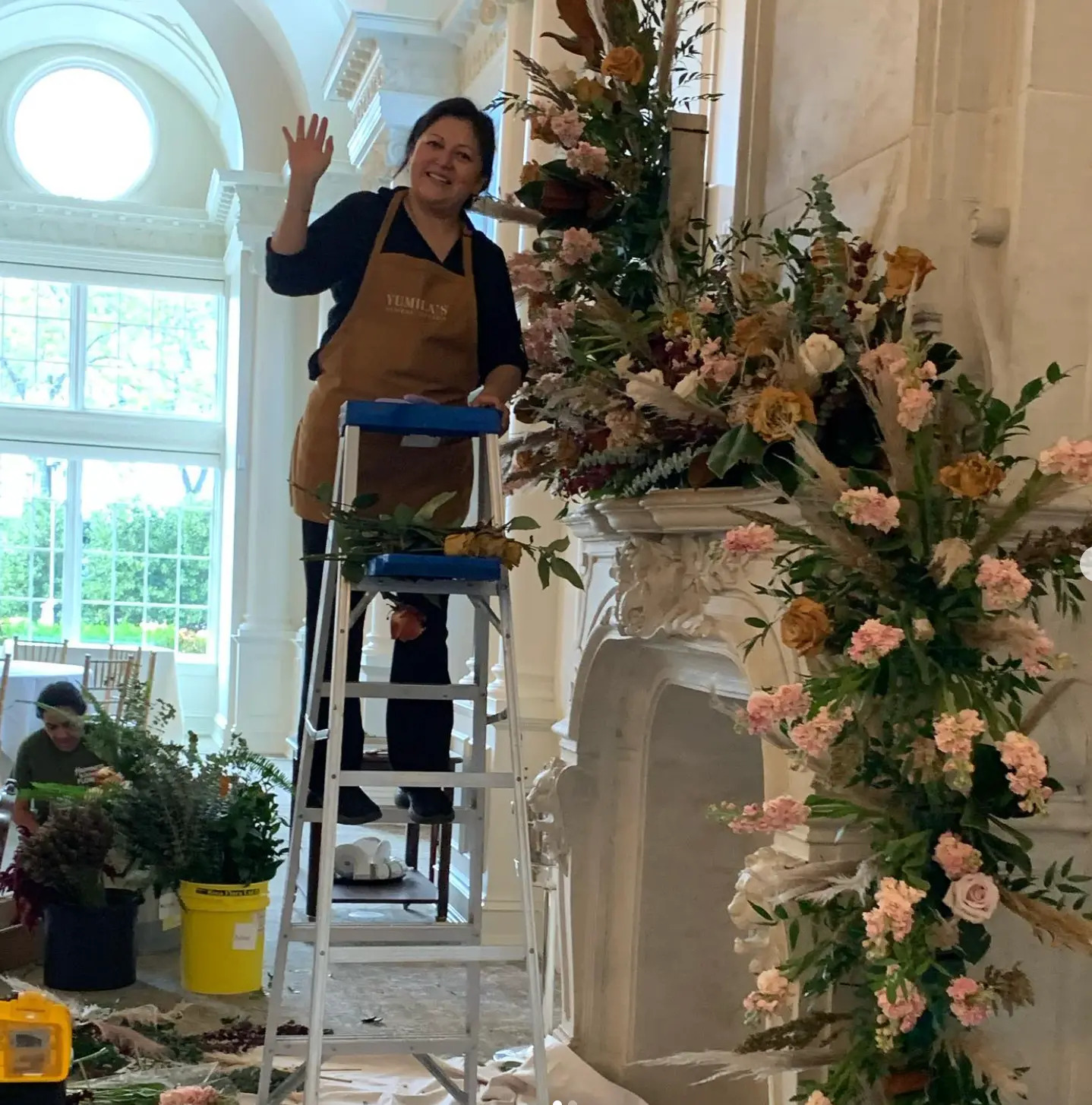 Yumila on a ladder installing a lush blush and ivory floral arrangement on a grand ballroom fireplace