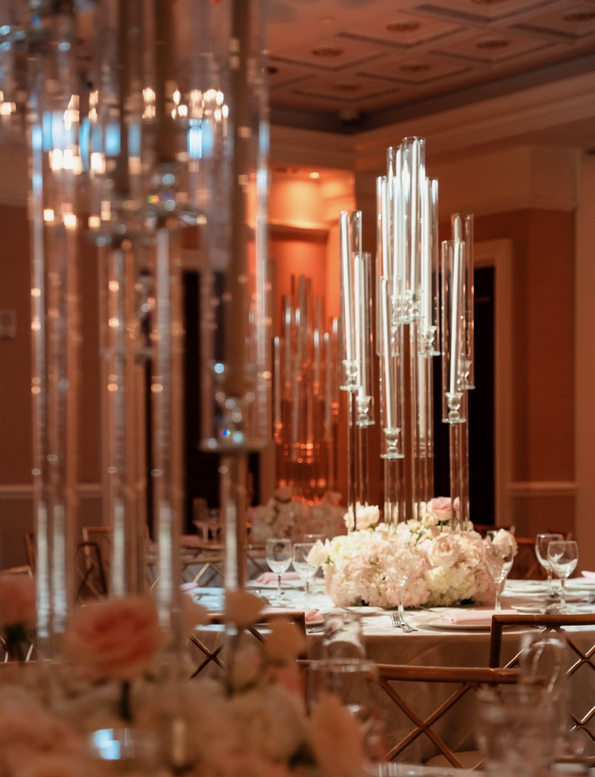 Tall acrylic candelabras above low blush rose and hydrangea centerpieces in a warmly lit ballroom