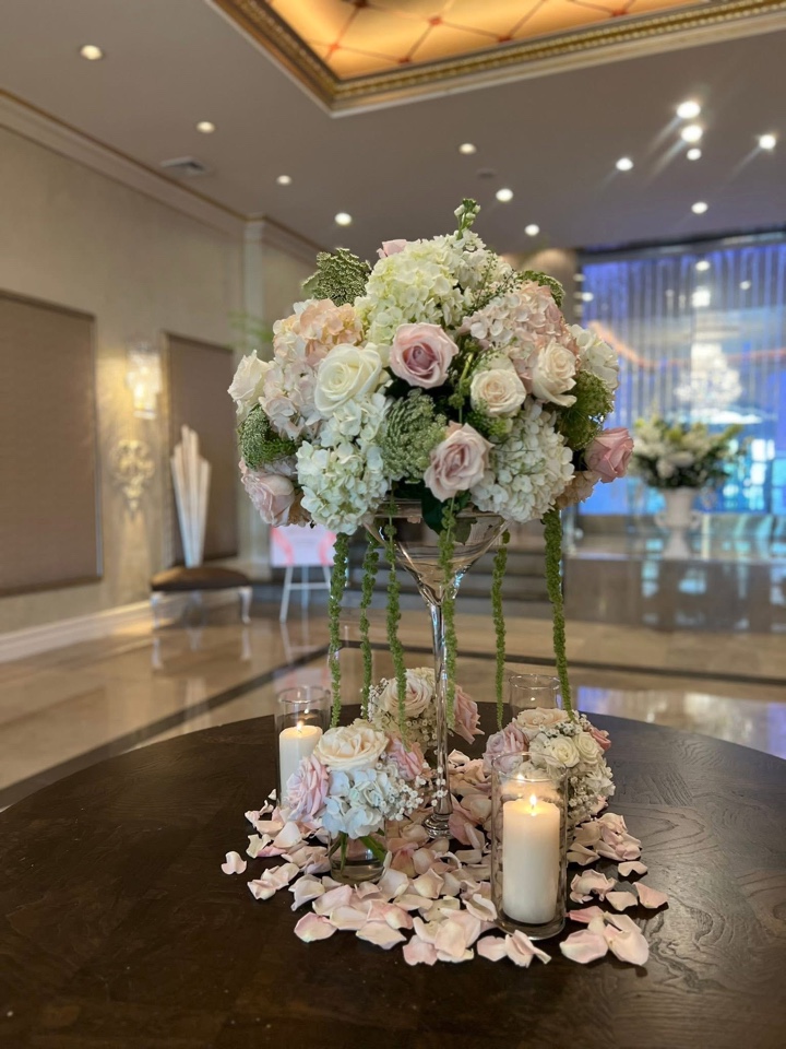 Tall martini-glass centerpiece of blush roses, white hydrangea and trailing amaranthus with candles and scattered petals