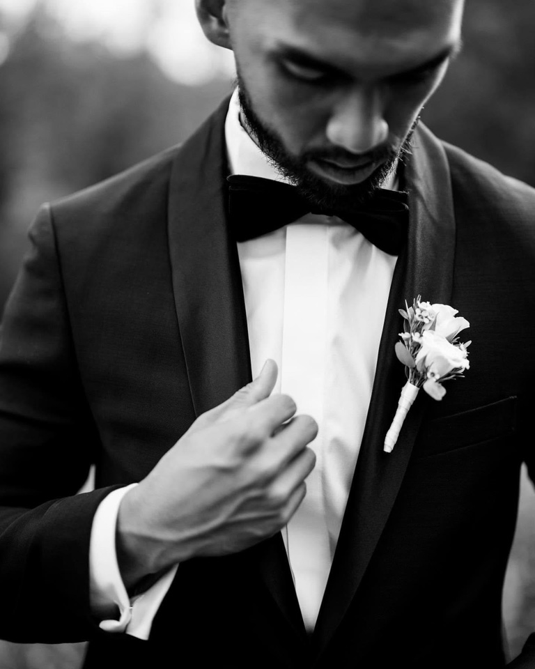 Black and white portrait of the groom adjusting his jacket, wearing a white rose boutonniere