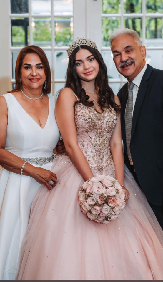 Birthday girl in a blush gown and tiara with her parents, holding a blush rose bouquet