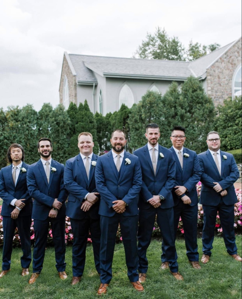 Groom and groomsmen in navy suits wearing white rose boutonnieres outside a stone chapel