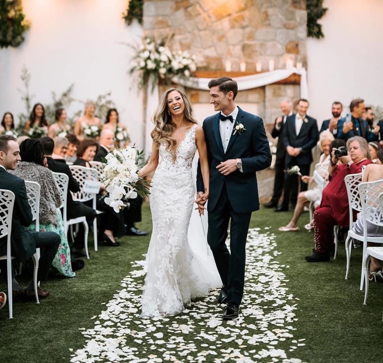 Newlyweds laughing down a white petal aisle with a white orchid and rose bouquet