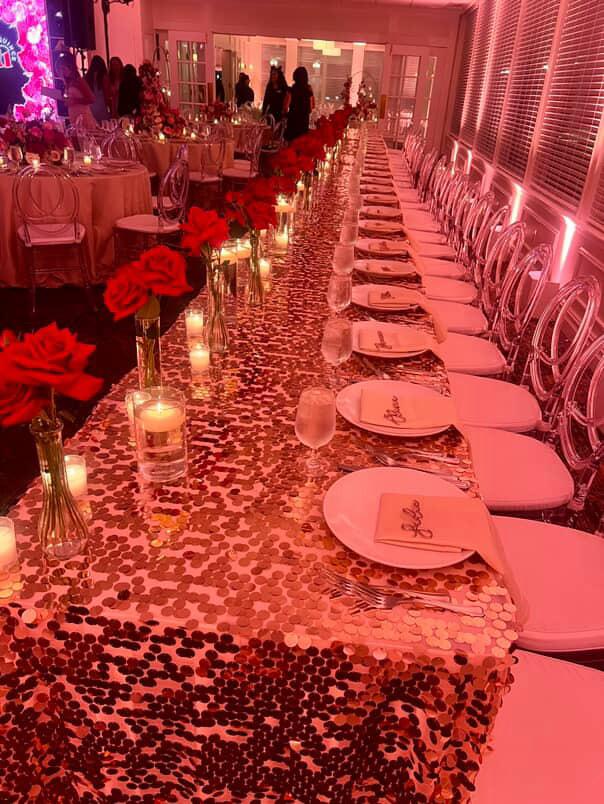 Long king's table in a rose-gold sequin linen with red rose bud vases, floating candles and ghost chairs