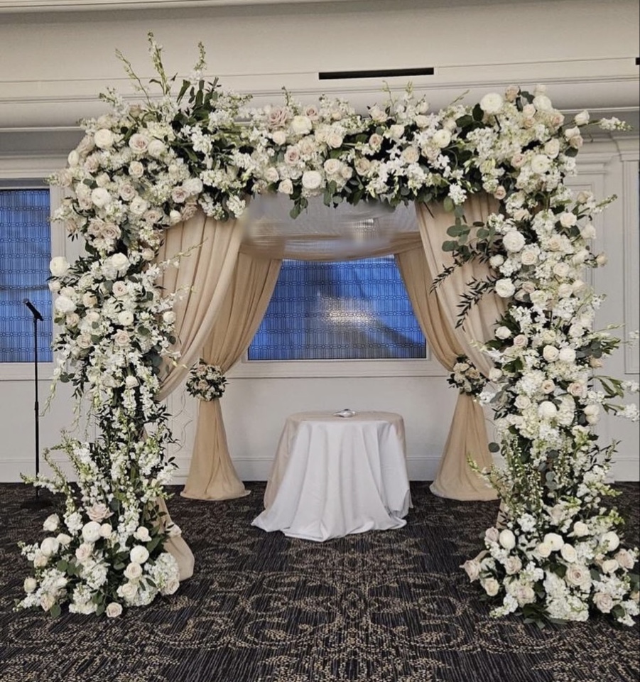 Square chuppah wrapped in white roses, stock and taupe draping with floral corners