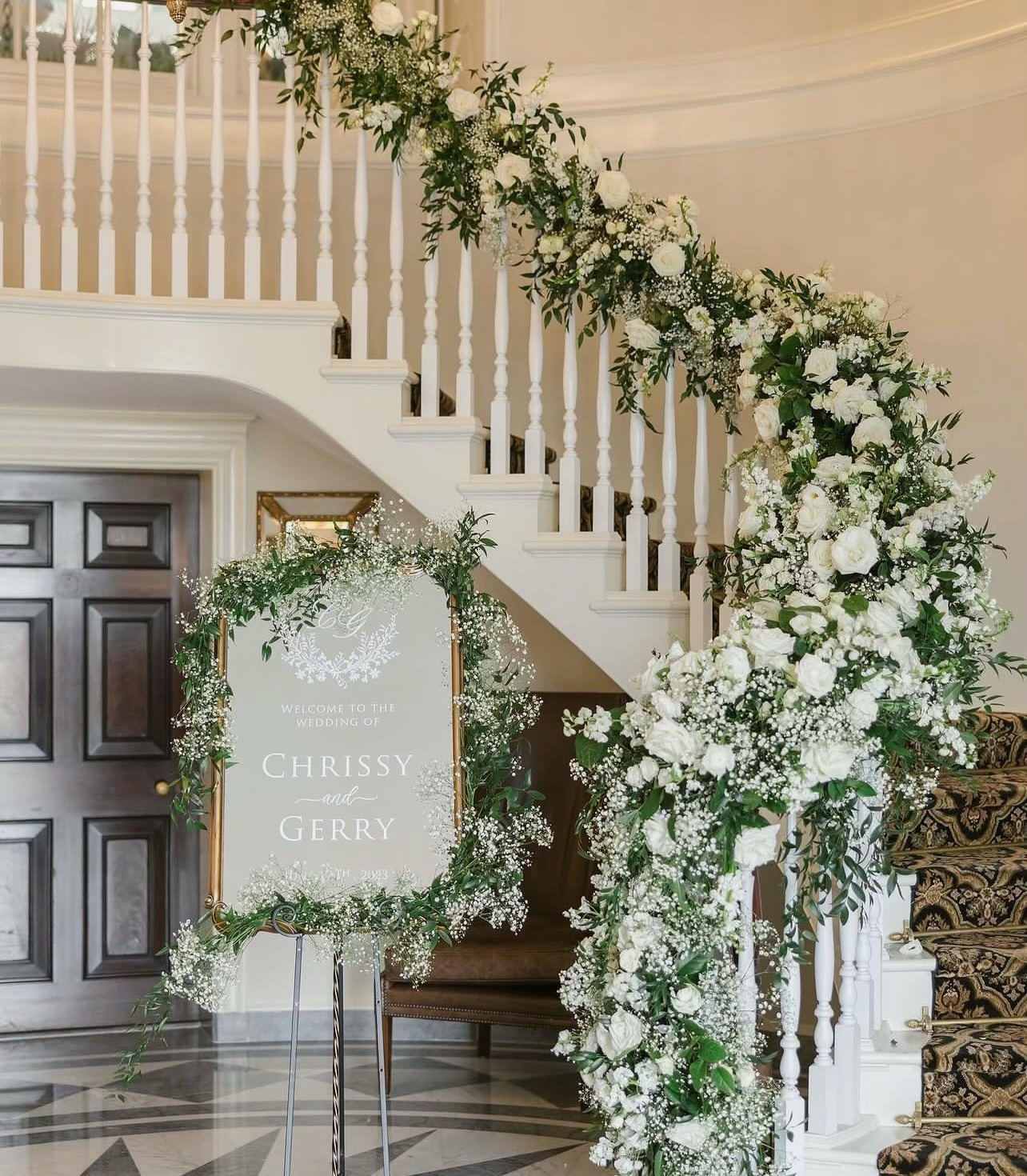 Estate staircase wrapped in white roses, baby's breath and greenery beside a garlanded welcome sign