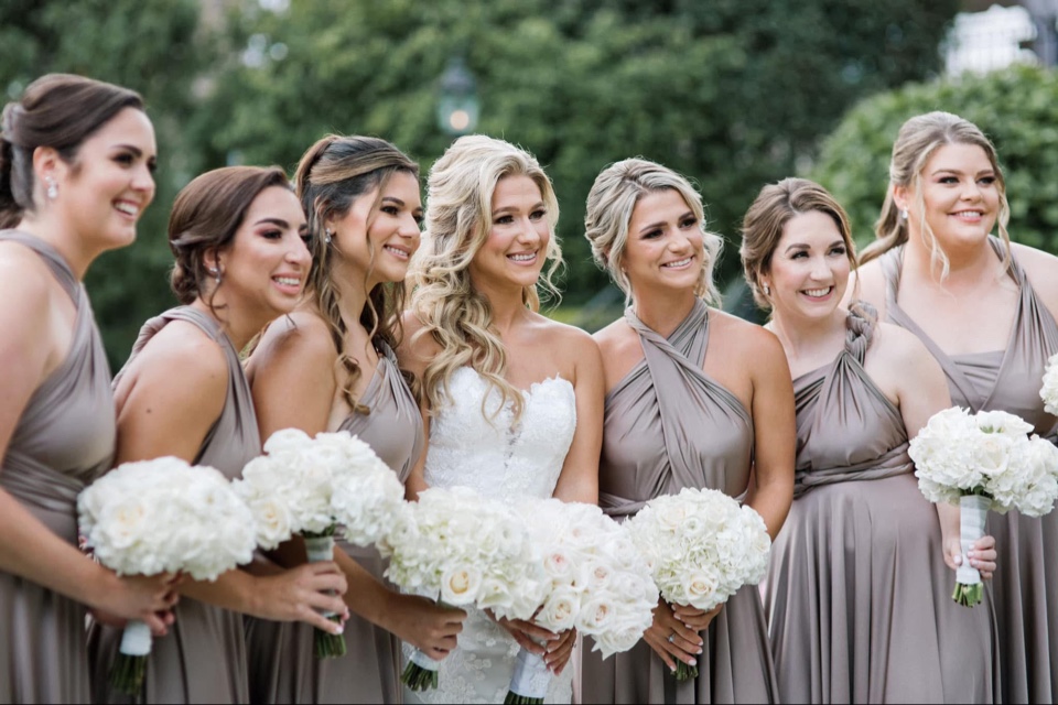 Bride laughing with bridesmaids in taupe gowns, each holding a white hydrangea and rose bouquet