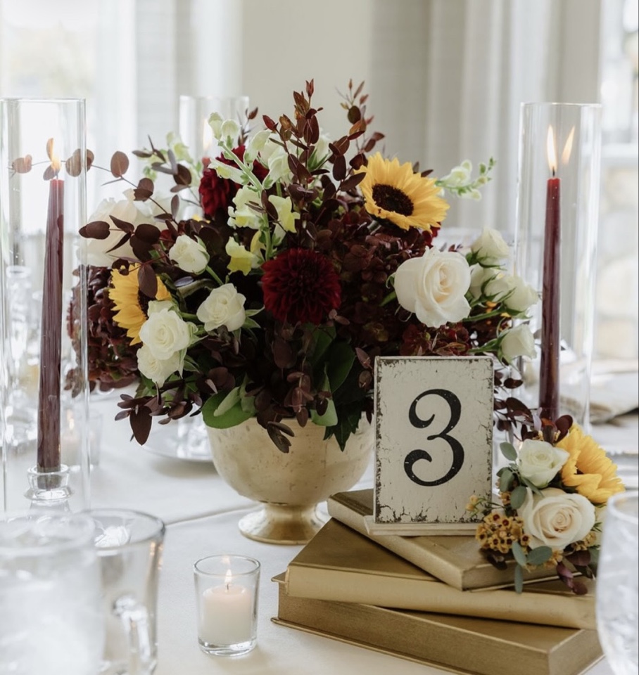 Fall centerpiece of sunflowers, burgundy dahlias and white roses in a gold compote with taper candles and a table number