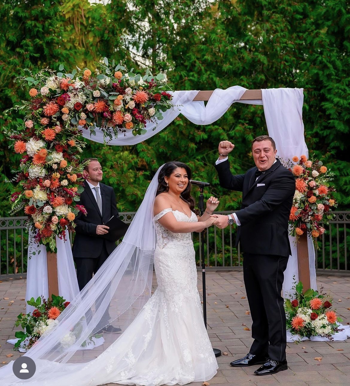 Couple celebrating beneath an arch of orange dahlias, burgundy roses and eucalyptus