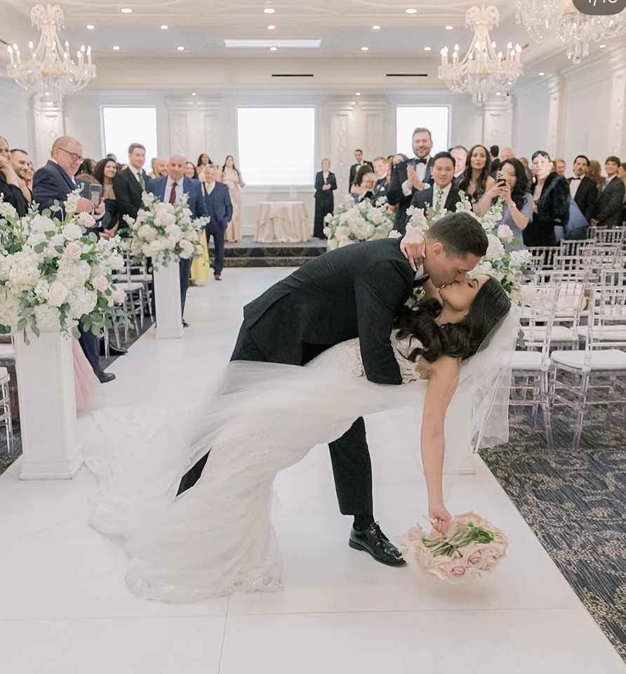 Groom dipping the bride for a kiss in a ballroom ceremony aisle lined with blush and white pedestal arrangements