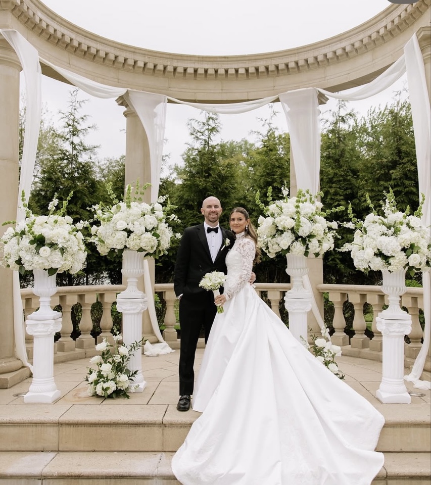Bride and groom beneath a circular stone rotunda draped in white fabric with tall white hydrangea and stock urns