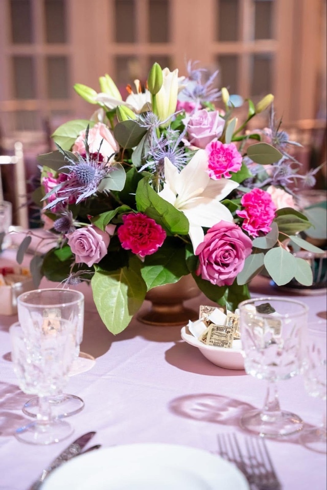 Low centerpiece of white lilies, fuchsia carnations, lavender roses and blue thistle on a lavender tablecloth