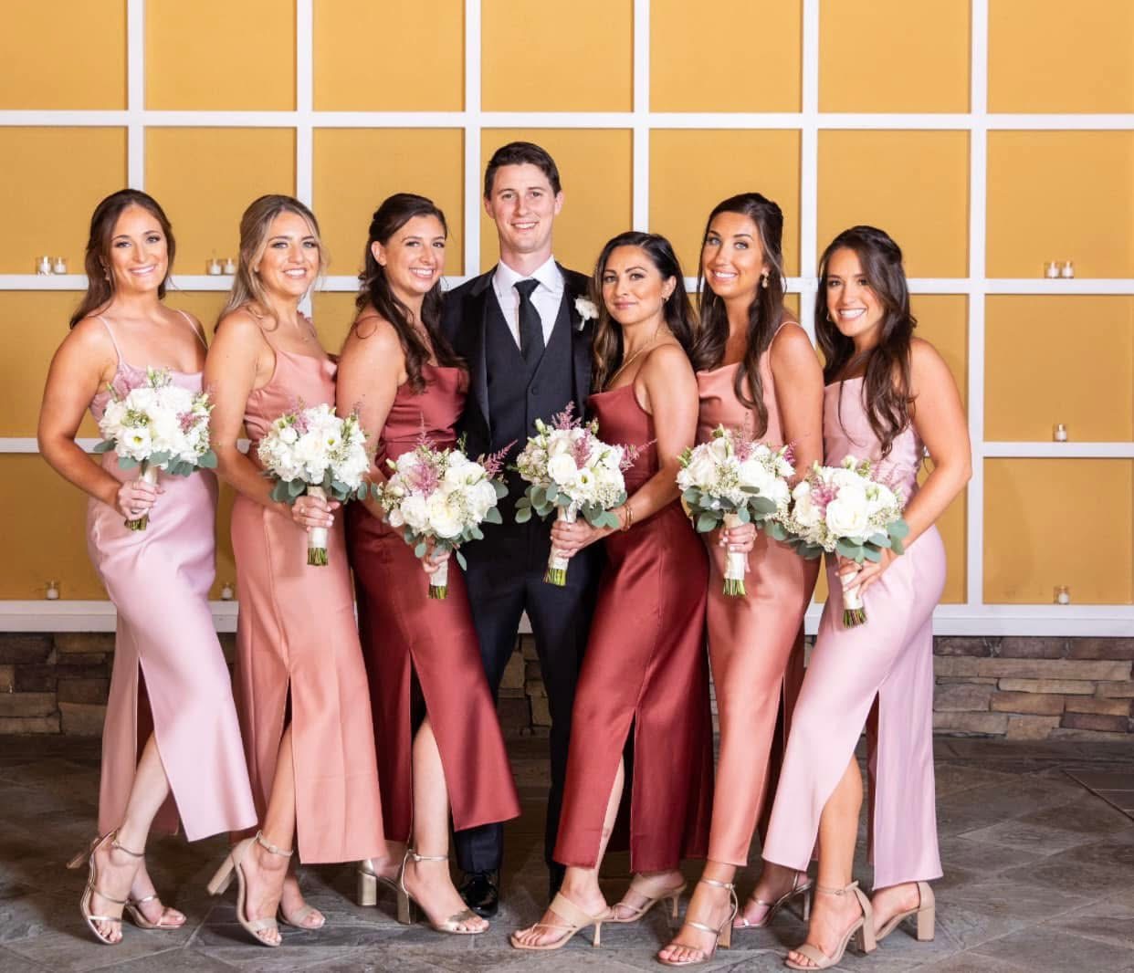 Bridesmaids in rose and terracotta gowns holding white rose and astilbe bouquets around the groom