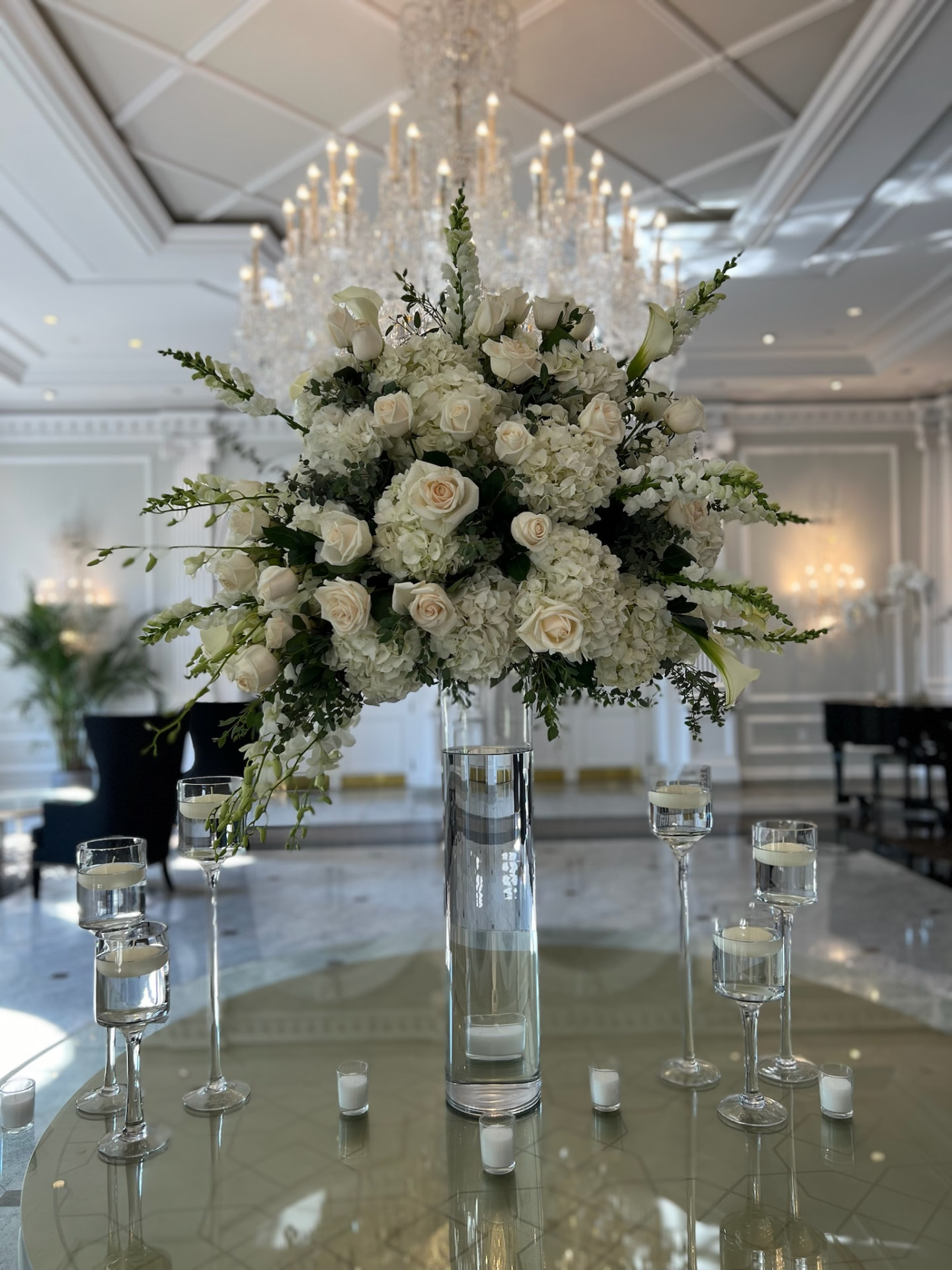 Tall white rose, hydrangea and orchid centerpiece on a glass pedestal beneath a crystal ballroom chandelier