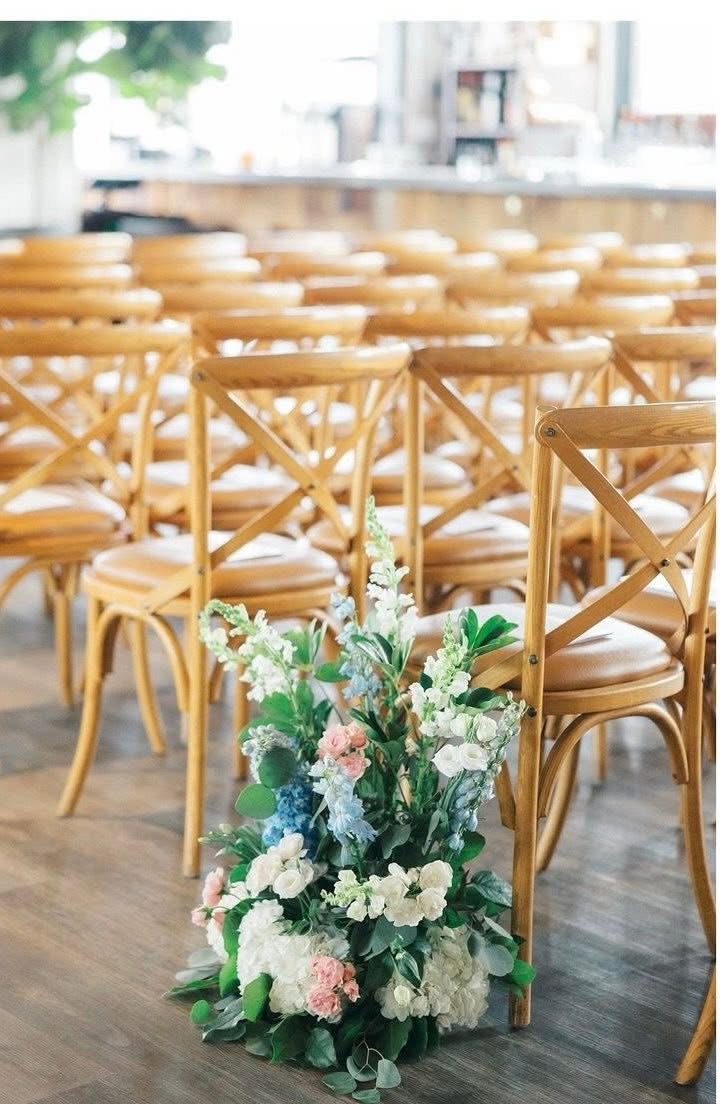 Cluster of blush roses, white hydrangea and blue delphinium beside wooden cross-back chairs on the ceremony aisle