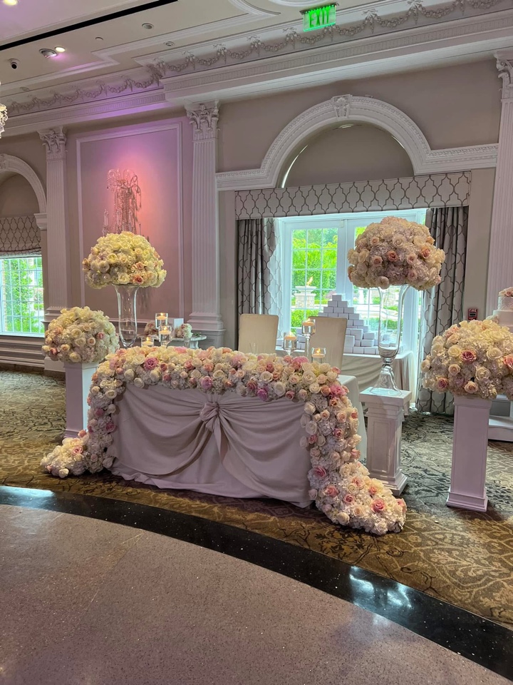 Sweetheart table swagged in fabric and covered in pink and white roses beside tall gold urn arrangements