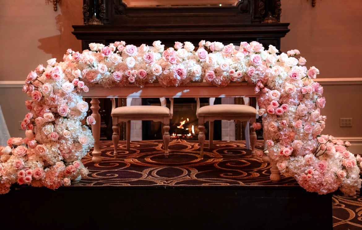 Sweetheart table framed by a full arch of blush and ivory roses in front of a lit fireplace