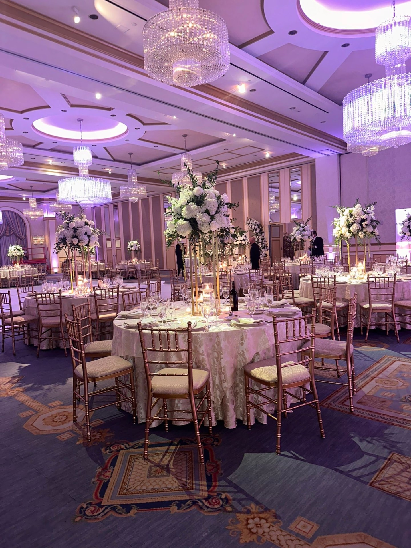 Ballroom set with tall white hydrangea and rose centerpieces on gold stands beneath crystal chandeliers