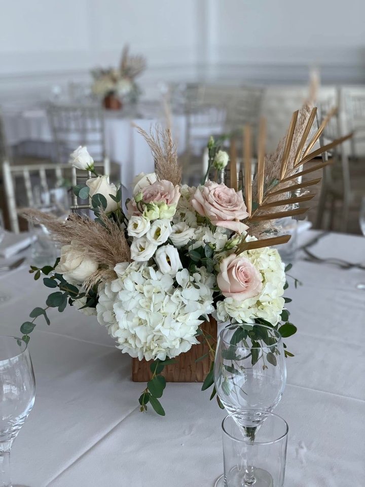 Wood box centerpiece of blush roses, white hydrangea, pampas grass and gilded palm leaves in a bright ballroom