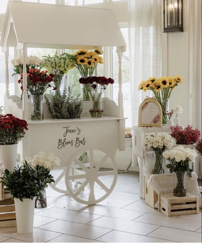 White flower cart labeled Jenni's Bloom Bar stocked with buckets of sunflowers, red and white blooms