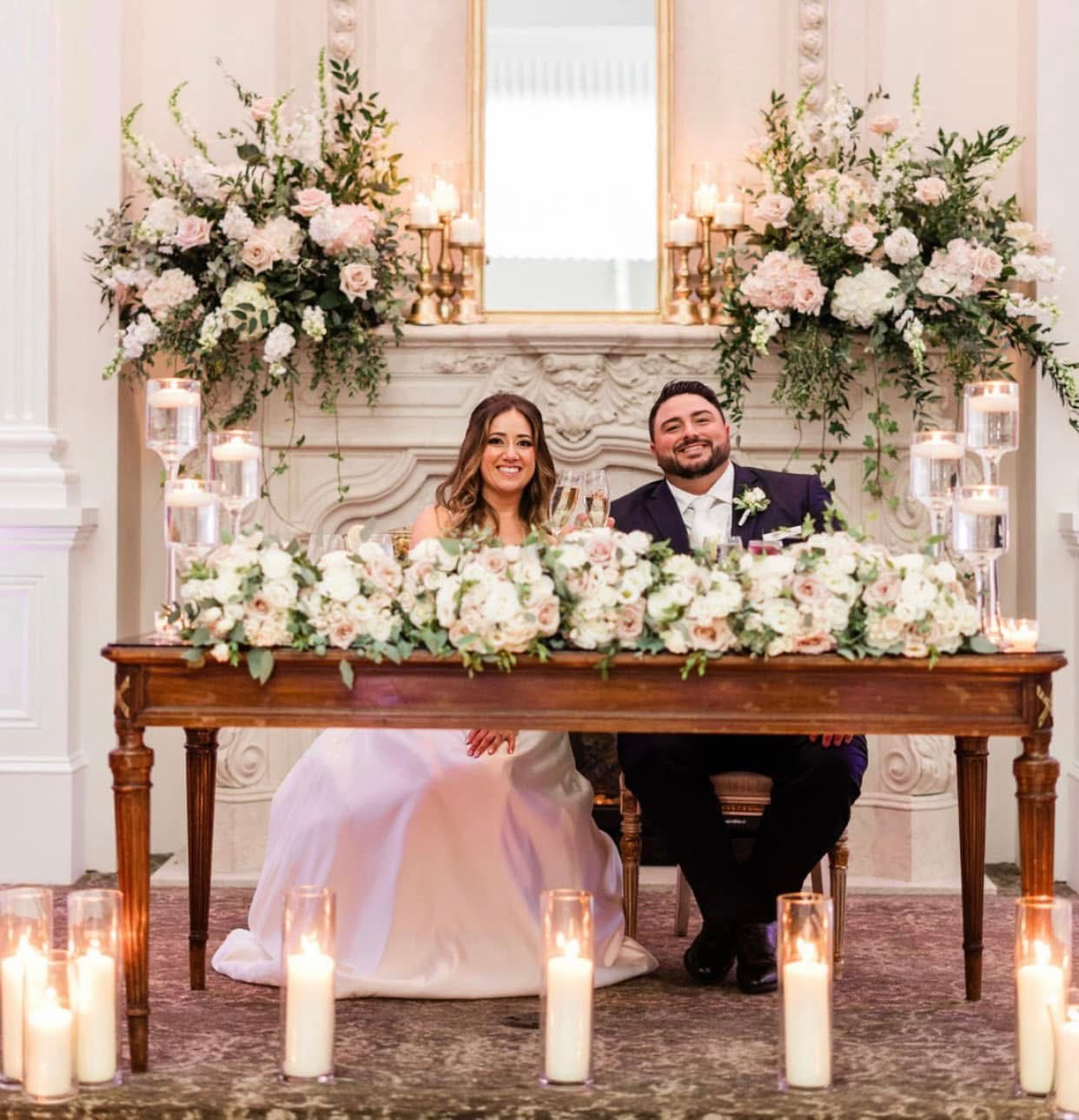 Couple at a sweetheart table crowned in blush and ivory blooms before a marble fireplace with candles