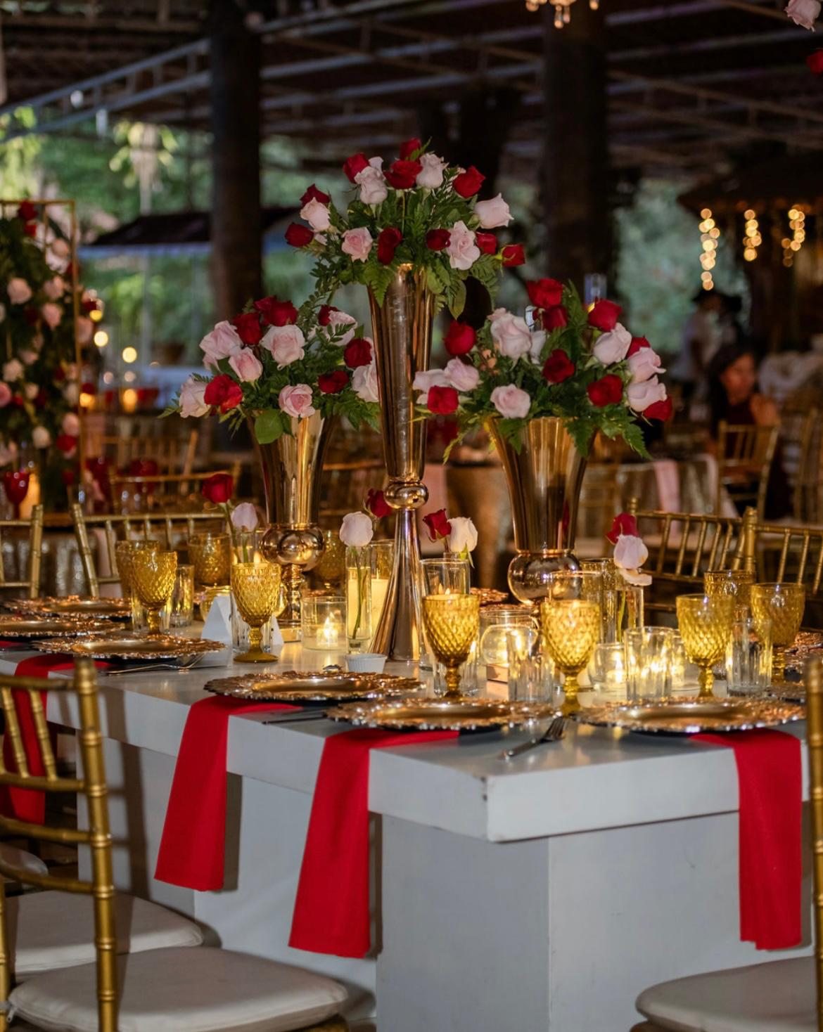 Head table crowned with three gold trumpet vases of red and blush roses above gold goblets and red runners