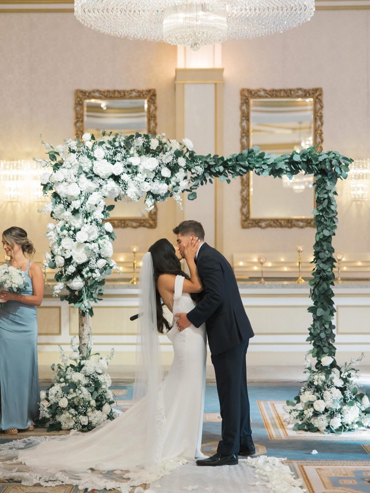 Couple kissing under a chuppah of white hydrangea, roses and trailing eucalyptus