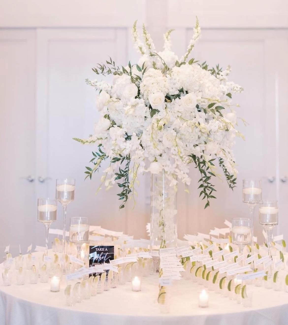 Escort card table with shot glass favors, lime wedges, and a tall white hydrangea centerpiece