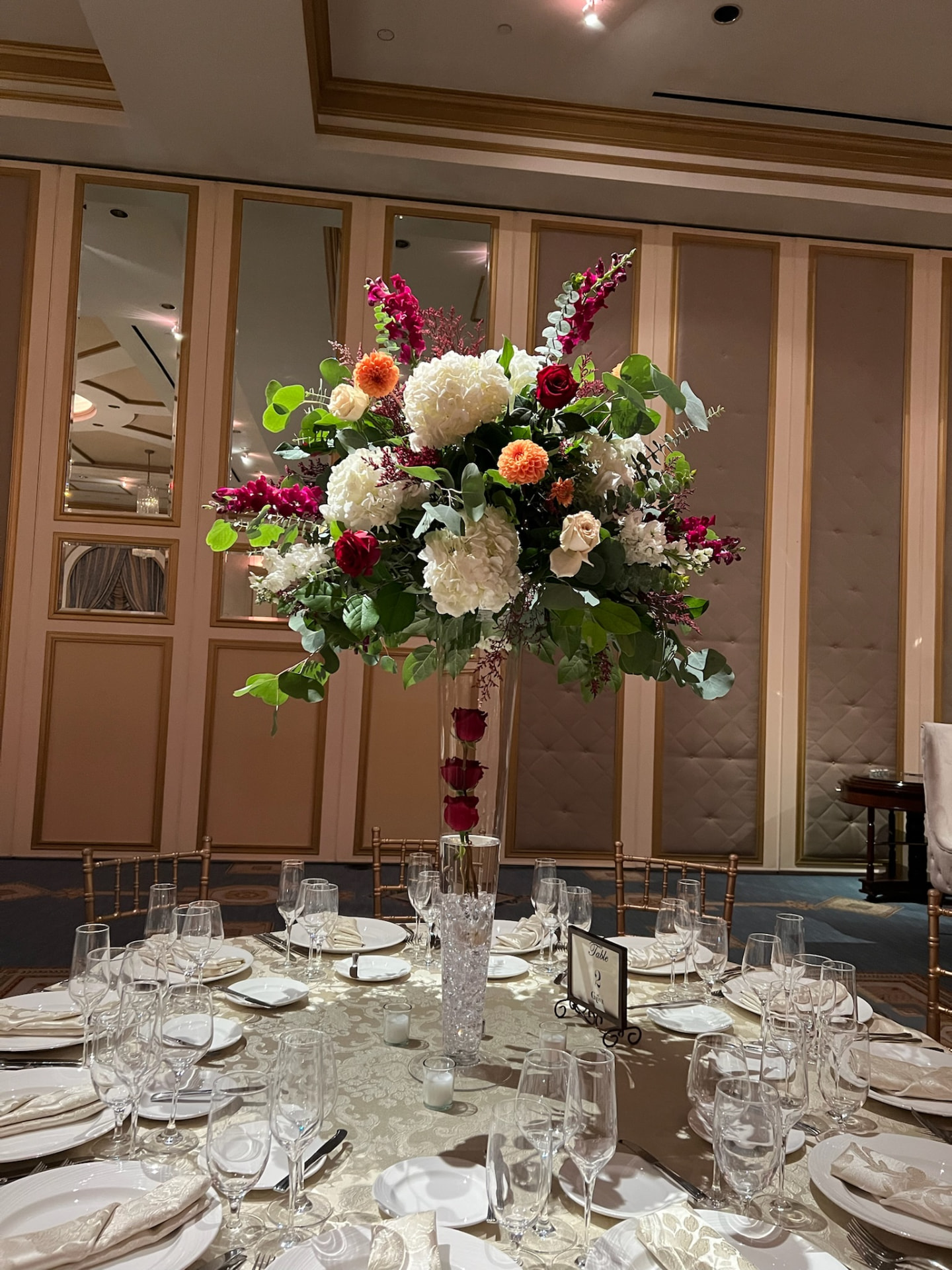 Tall centerpiece of white hydrangea, red roses, orange dahlias and magenta orchids over a formal place setting