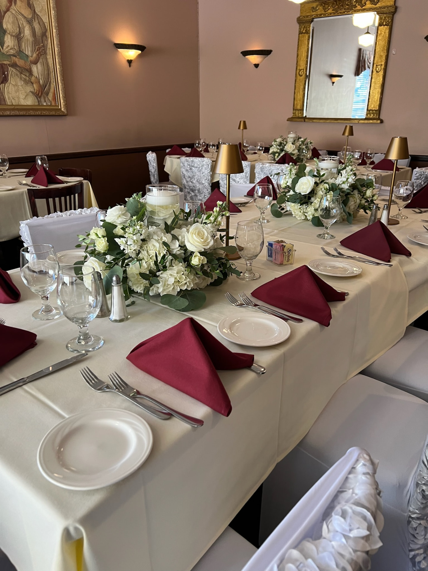 Long banquet table with white rose and eucalyptus runners, burgundy napkins and brass table lamps