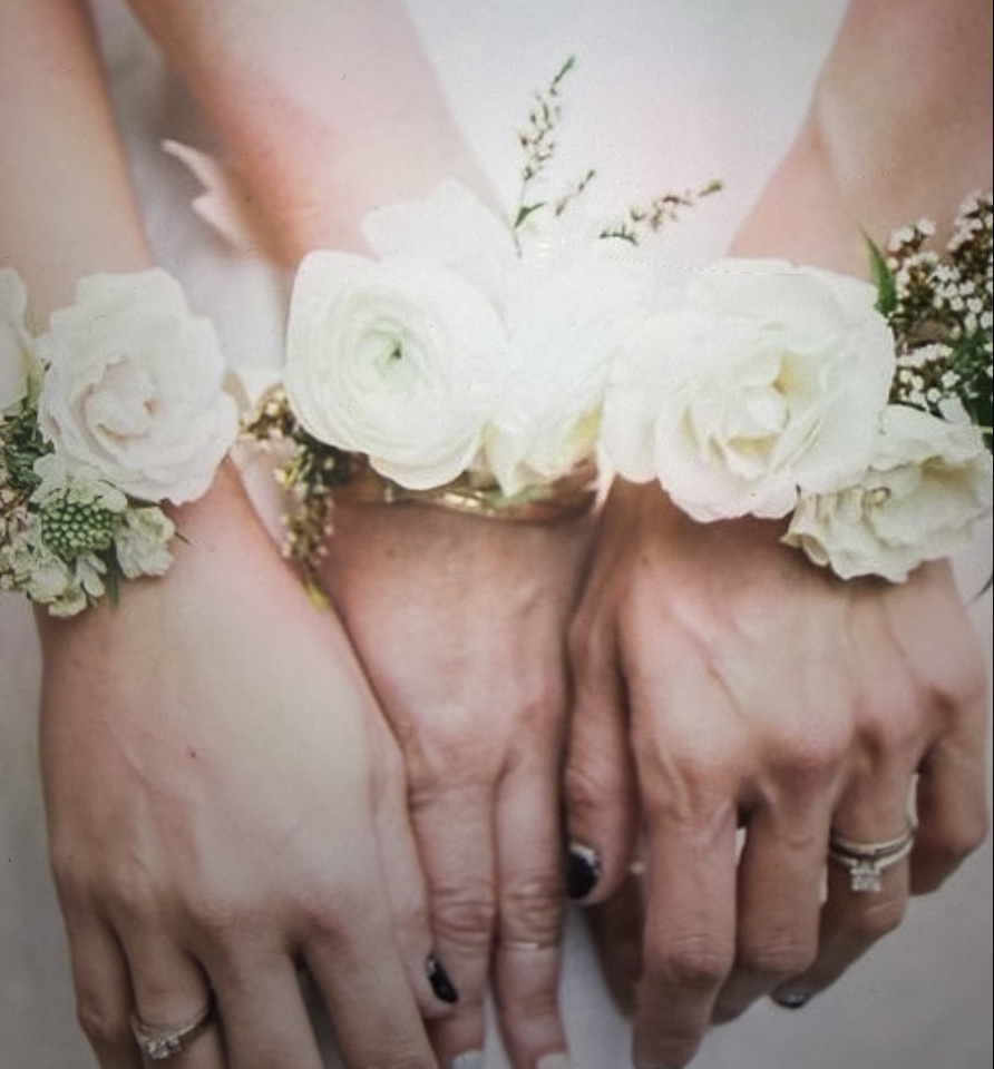Three wrists wearing white ranunculus and rose corsages on gold cuffs