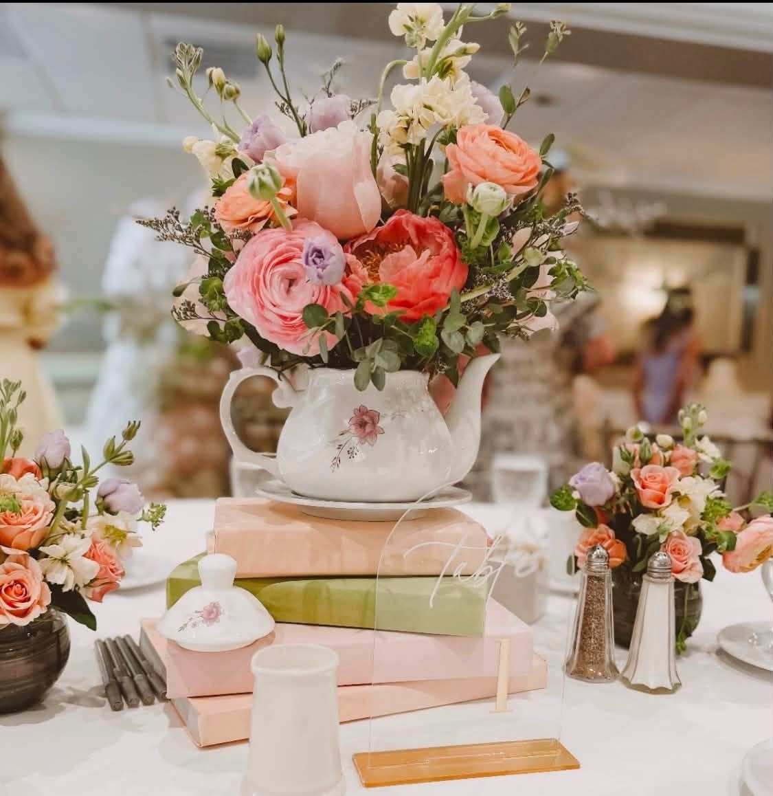 Tea party table with a teapot centerpiece of pink peonies and ranunculus on pastel books, flanked by rose posies