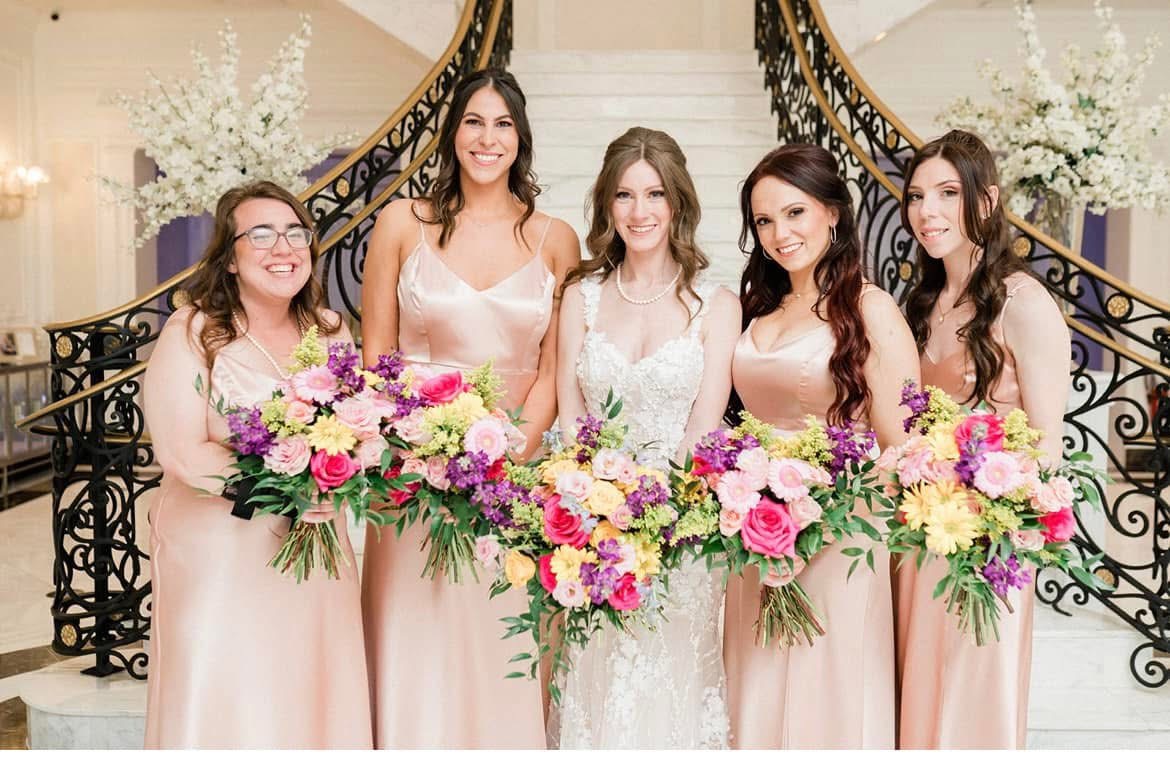 Bride and bridesmaids in blush satin holding bright bouquets of hot pink roses, yellow daisies and purple stock