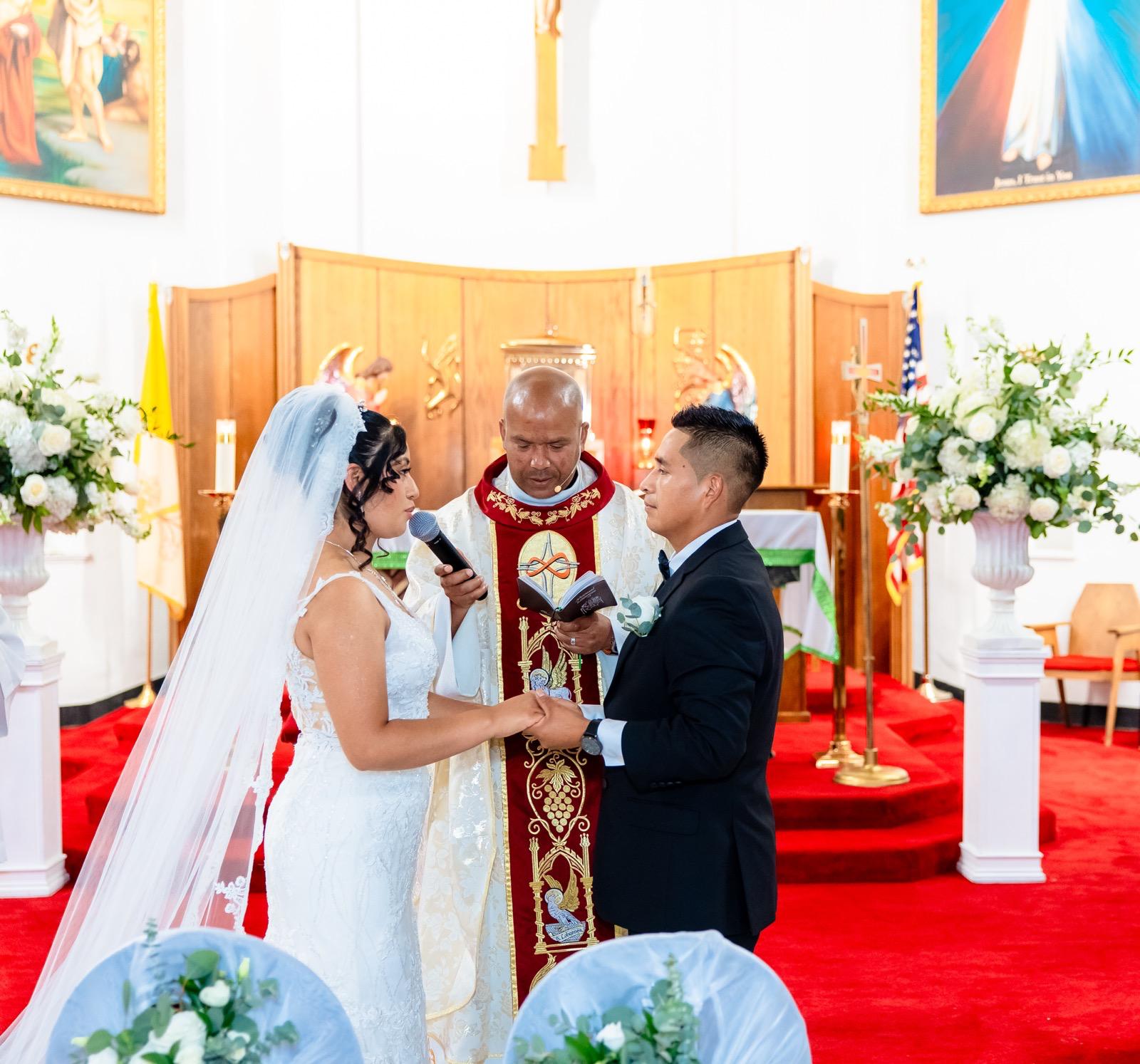 Couple exchanging vows at the church altar between large white floral arrangements