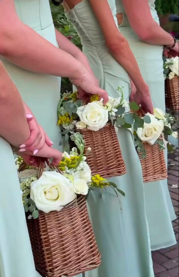 Bridesmaids in sage green dresses holding wicker baskets of white roses and garden accents