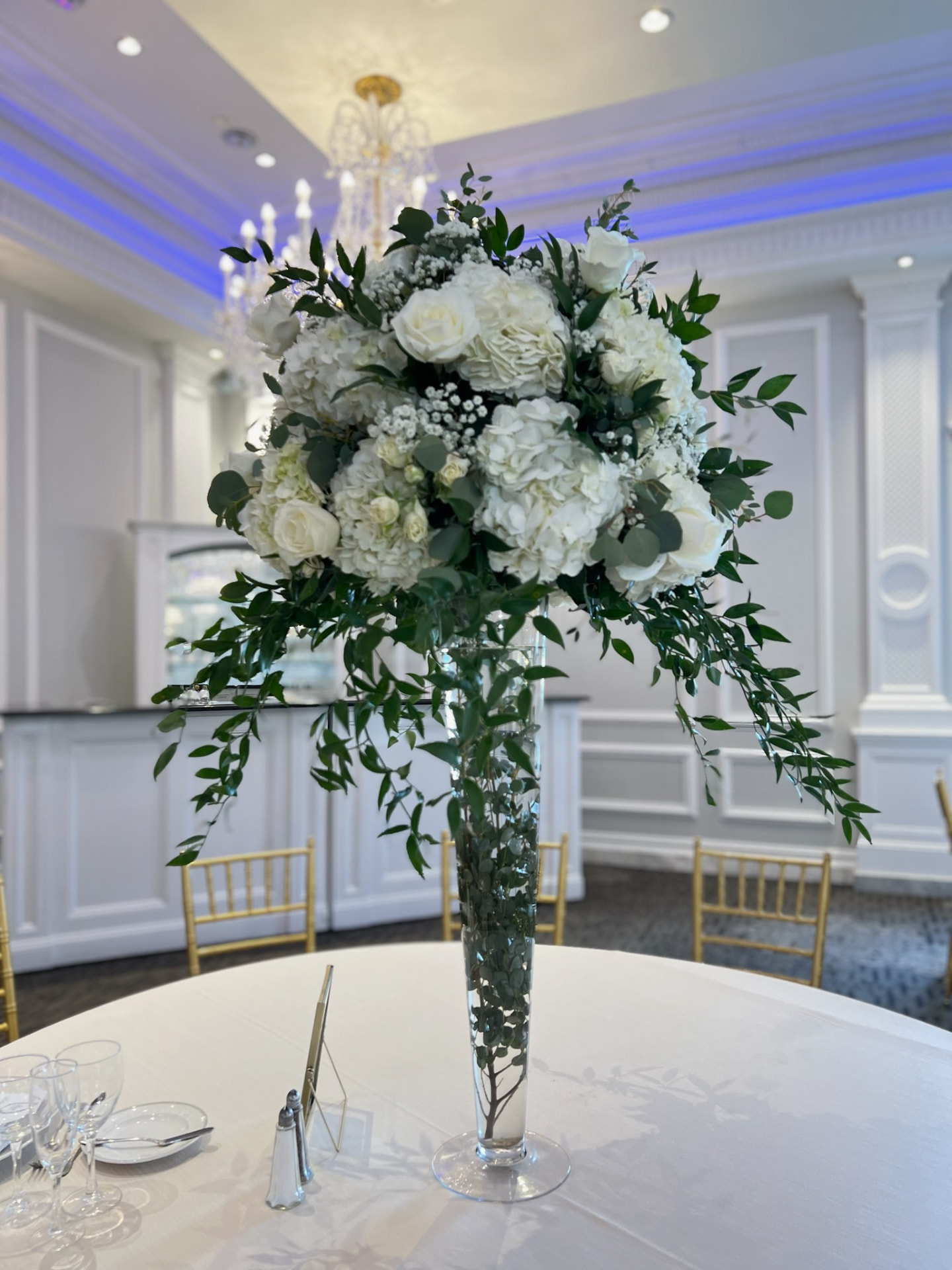 Tall white hydrangea, rose and trailing greenery centerpiece in a glass pilsner vase in a blue-lit ballroom