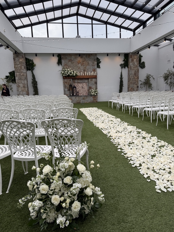 Atrium ceremony under a glass ceiling with a white rose petal aisle and a low hydrangea arrangement